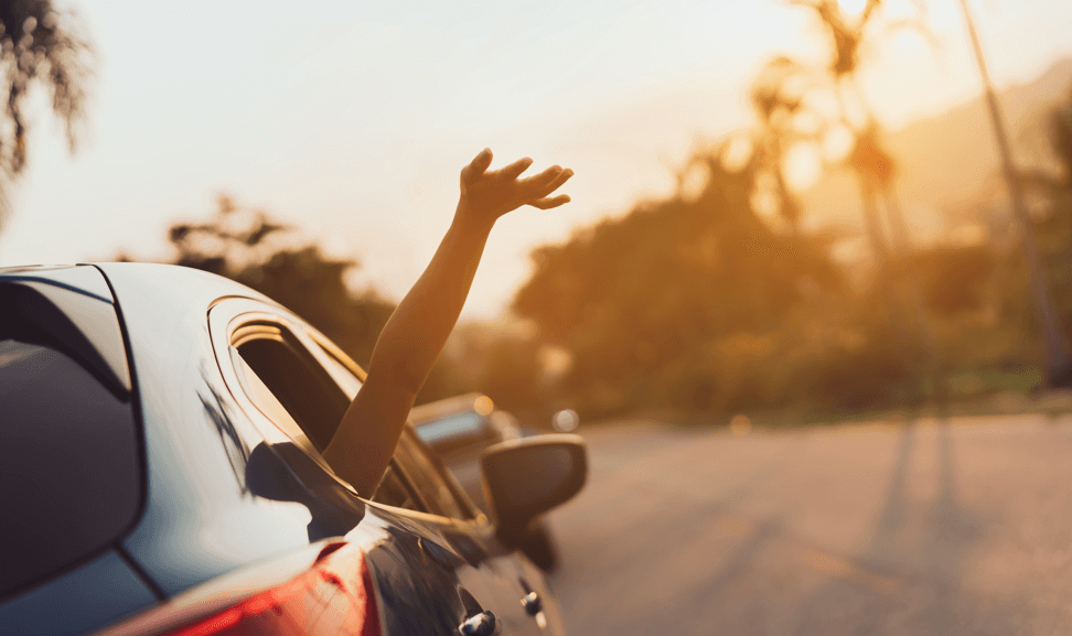 Voiture de location en mouvement dans laquelle un passager sort le bras par la fenêtre avec un coucher de soleil en arrière fond